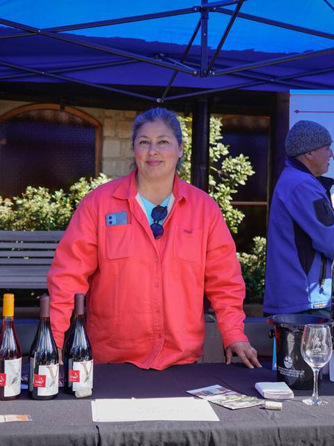 Una mujer sonriendo detrás de una mesa con botellas de vino en un evento al aire libre.