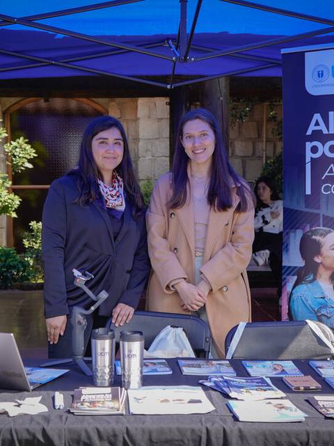 Dos mujeres posando frente a un stand informativo de educación.