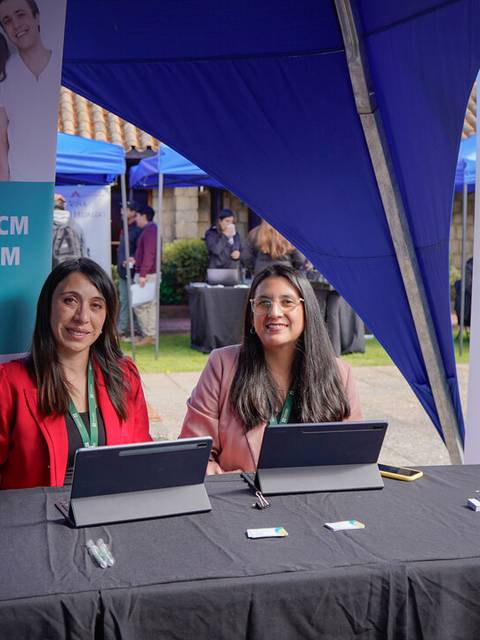 Dos mujeres sonrientes están sentadas en un stand con información sobre la Clínica UCM durante un evento al aire libre.