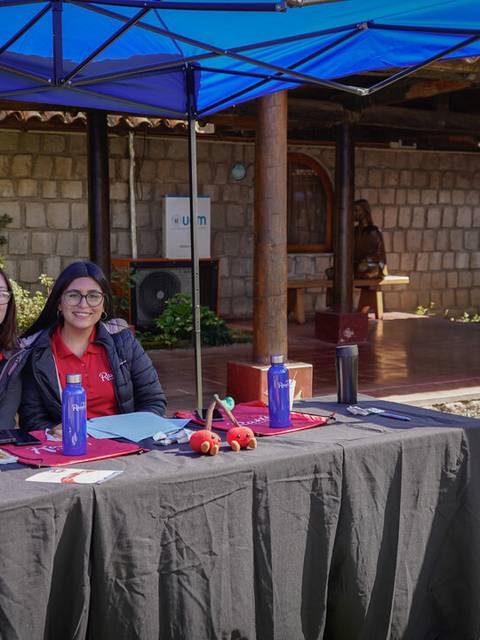 Dos mujeres sonrientes están sentadas en una mesa bajo una carpa, con un computador y carteles de una empresa de cerezas.