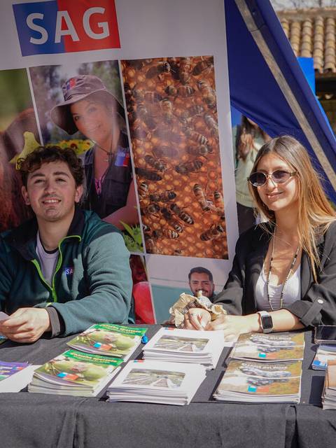 Dos jóvenes se encuentran en un stand al aire libre, mostrando material informativo.
