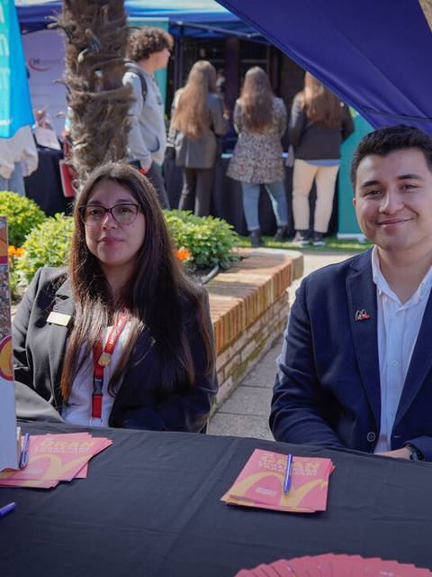 Dos jóvenes se encuentran sentados en una mesa frente a un cartel de McDonald's durante un evento al aire libre.