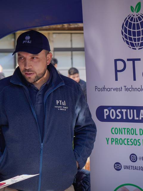 Una persona que está de pie al lado de un cartel sobre tecnología y procesos en postcosecha, conversando con otras personas.