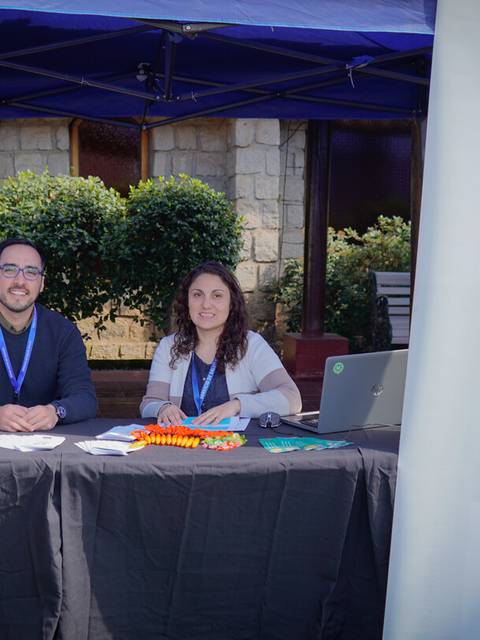 Dos personas están sentadas detrás de una mesa en un evento al aire libre, con un banner del Servicio de Salud Maule.