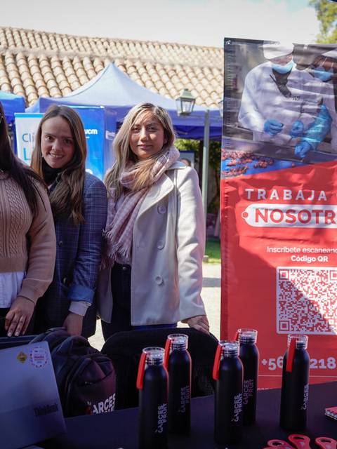 Tres mujeres posan sonrientes frente a un stand en un evento al aire libre.