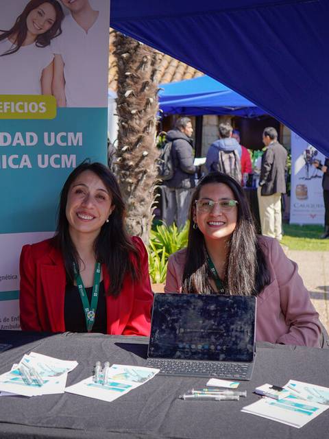 Dos mujeres sonrientes están sentadas detrás de una mesa en un evento, promocionando una clínica con materiales informativos.
