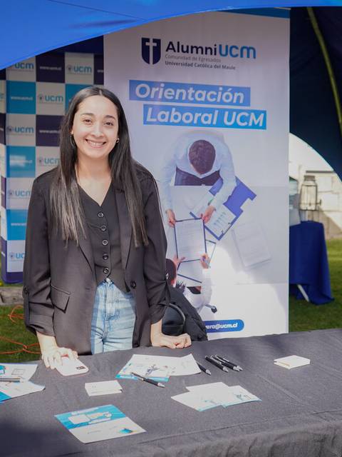 Una mujer sonriente está de pie frente a una mesa con materiales informativos sobre orientación laboral en un evento.