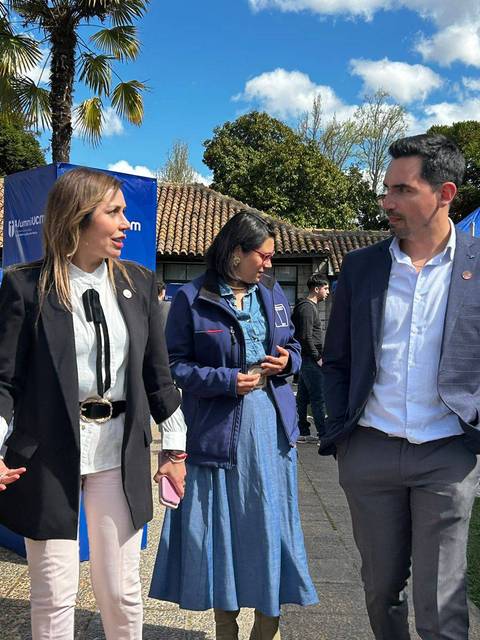 Tres personas conversan en un evento al aire libre con stands y palmeras de fondo.