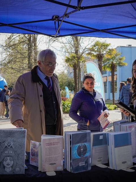 Un grupo de personas se encuentra en un evento al aire libre, observando libros expuestos en una mesa bajo un toldo azul.
