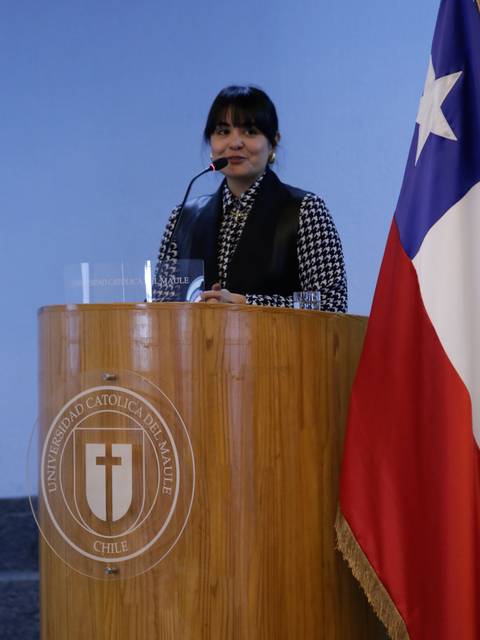 Una mujer presenta en un podio con el logo de la Universidad Católica del Maule y una bandera chilena al lado.