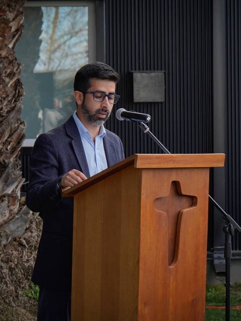 Un hombre habla desde un podio frente a una mesa adornada con flores y un símbolo religioso.