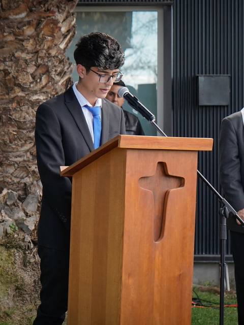 Un joven vestido con traje habla frente a un podio con una cruz, mientras un sacerdote lo observa en un evento al aire libre.