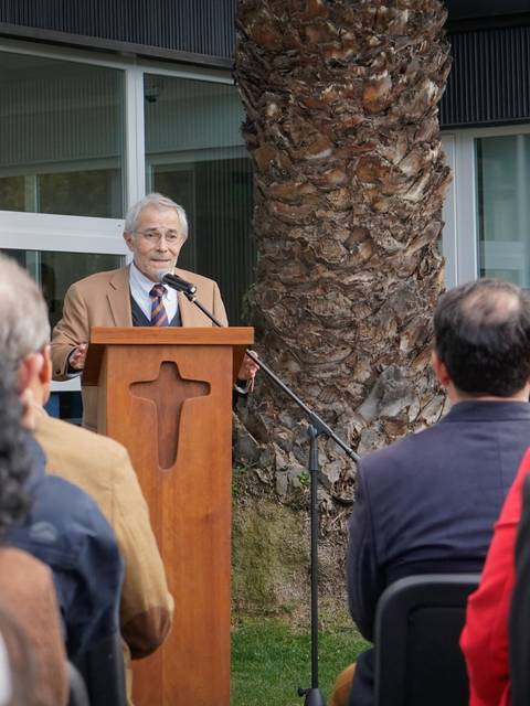 Un hombre mayor está hablando desde un podio en un evento al aire libre, con una audiencia de pie frente a él.