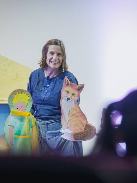 Una mujer sostiene dos figuras de papel, una de un niño y otra de un zorro, mientras sonríe frente a una audiencia.