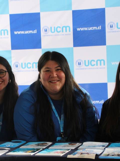 Tres mujeres sonrientes posando frente a un stand con el logo de UCM.