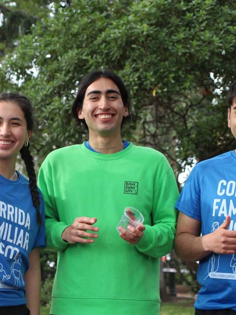 Un grupo de tres jóvenes sonrientes en un evento al aire libre, vestidos con camisetas de color azul y verde.