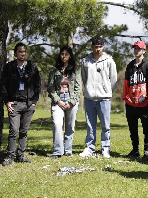 Un grupo de jóvenes escucha atentamente a un instructor bajo un árbol en un entorno natural.