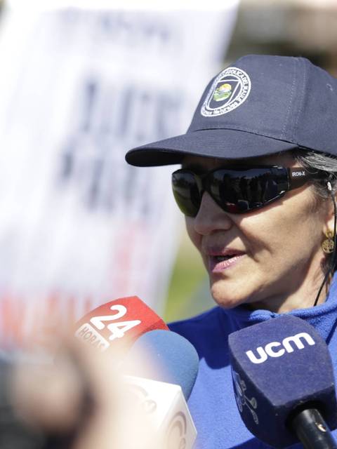 Una mujer con gafas de sol y gorra azul habla ante varios micrófonos en un evento al aire libre.