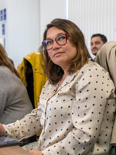 Una mujer con gafas de vista, vestida con una blusa blanca con puntos negros, observa atentamente en un entorno de reunión.