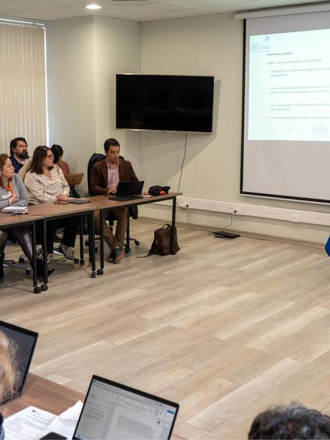Una reunión en un aula con varias personas sentadas frente a un presentador que está hablando.