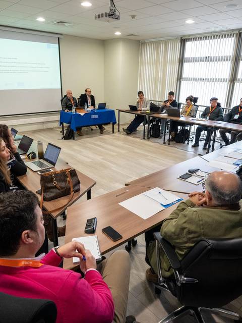 Una reunión en una sala con varias personas sentadas alrededor de una mesa en forma de U.