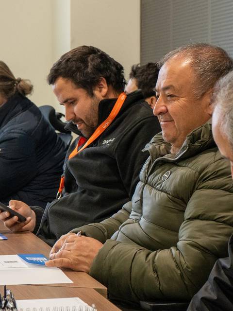 Un grupo de personas sentadas en una sala de reuniones, algunas mirando sus dispositivos móviles y otras prestando atención.