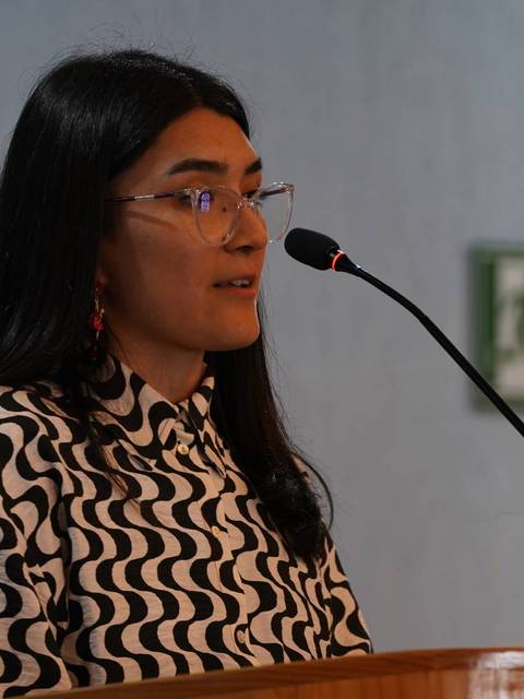 Una mujer con gafas y pelo largo habla desde un atril en un evento.