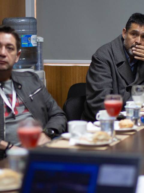 Dos hombres se encuentran sentados en una mesa durante una reunión, rodeados de bebidas y alimentos.
