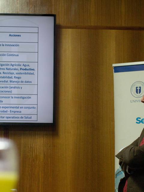 Un hombre observa una presentación en una sala de conferencias mientras se muestra una tabla con información variada en una pantalla.