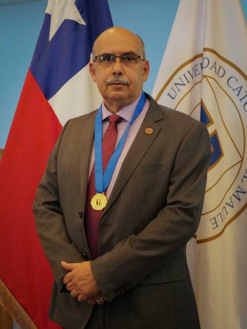 Un hombre con medalla dorada, vestido formalmente, posa frente a una bandera chilena y una insignia de universidad.