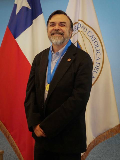 Un hombre sonriente de pie frente a banderas chilena y de la universidad.