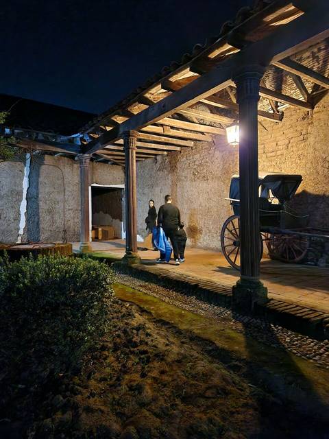 Una escena nocturna en un patio con una carretilla antigua y personas conversando bajo la luz de una lámpara.