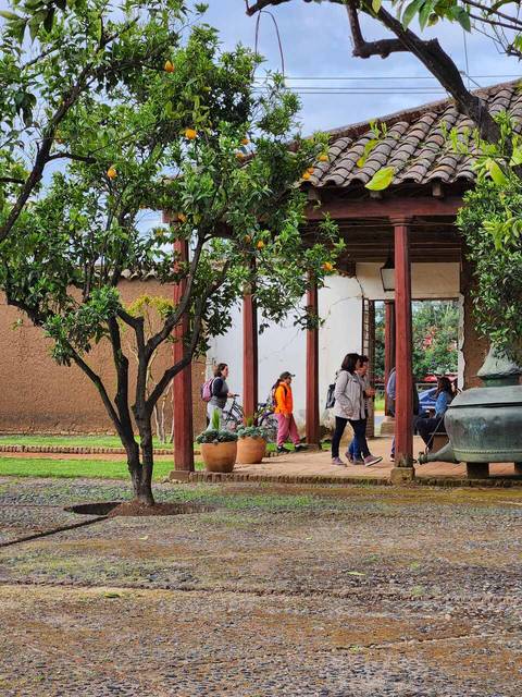 Un grupo de personas camina entre árboles y plantas en un entorno al aire libre con una construcción de estilo tradicional al fondo.