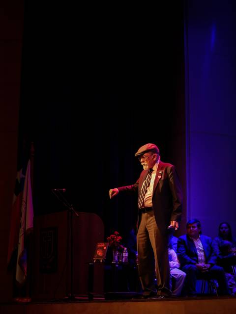 Una persona mayor habla en un escenario con un ambiente iluminado y un público atento.