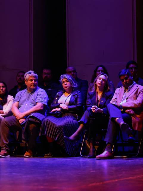 Un grupo de personas sentadas en un escenario iluminado de forma tenue, observando una presentación.
