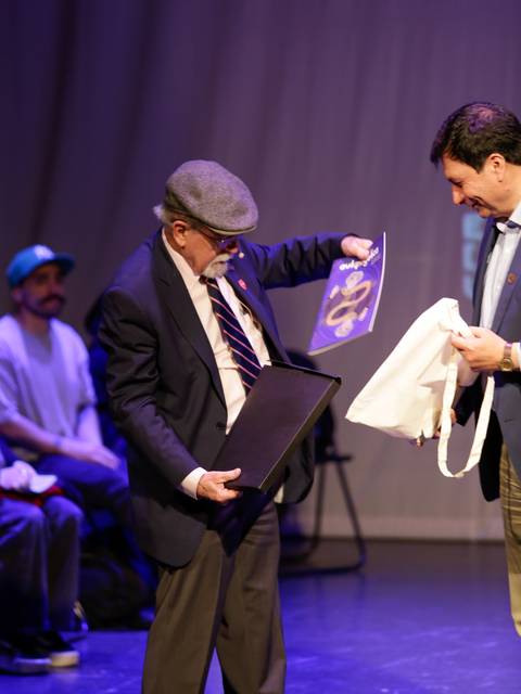 Un anciano recibe un reconocimiento en un evento mientras otro hombre le muestra un obsequio.