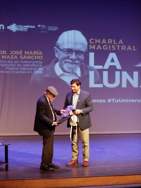 Un evento académico donde dos hombres intercambian un documento en el escenario.