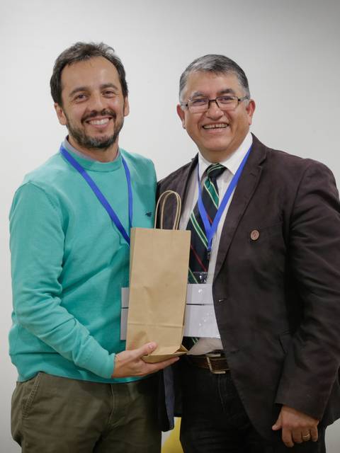 Dos hombres sonrientes posan juntos sosteniendo una bolsa de papel en un evento.