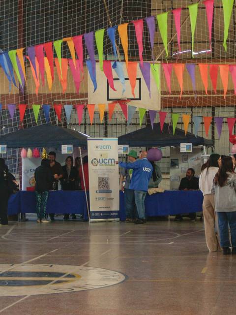 Un evento en un gimnasio con numerosos puestos decorados con banderines de colores y personas interactuando.