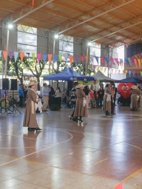 Una feria escolar con muchas personas y stands decorados con banderines de colores en un gimnasio.