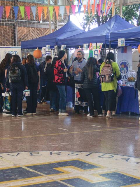 Una multitud de personas se encuentra en un evento dentro de un recinto deportivo, rodeadas de stands azules y decoraciones coloridas.