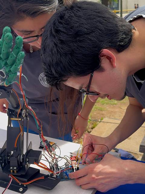 Dos estudiantes trabajan en un proyecto de robótica, manipulando un brazo robótico y cables.