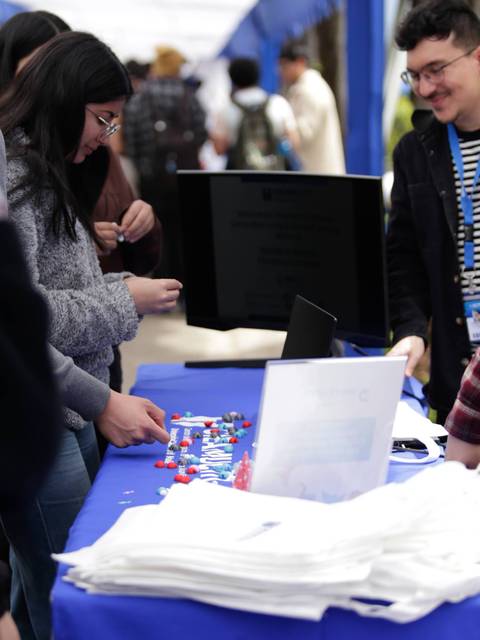Un grupo de personas interactúa en un evento al aire libre con mesas y material sobre una superficie azul.