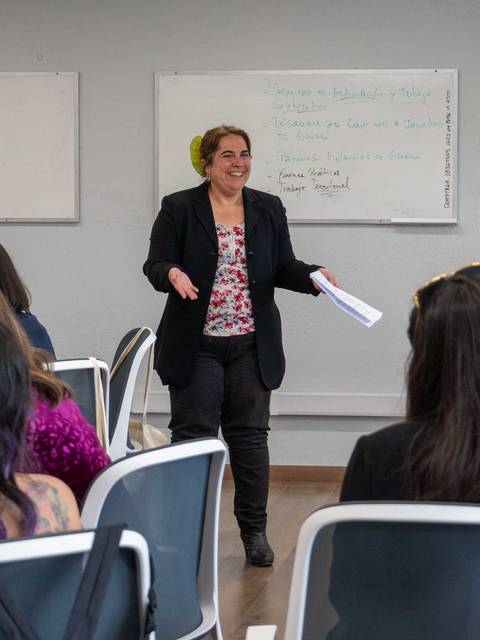 Una profesora está impartiendo una clase en un aula rodeada de estudiantes.
