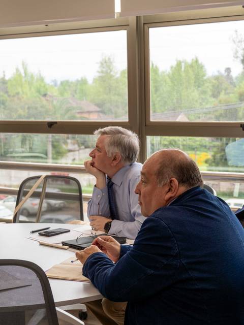 Dos hombres sentados en una mesa de trabajo, mirando hacia afuera a través de una ventana.