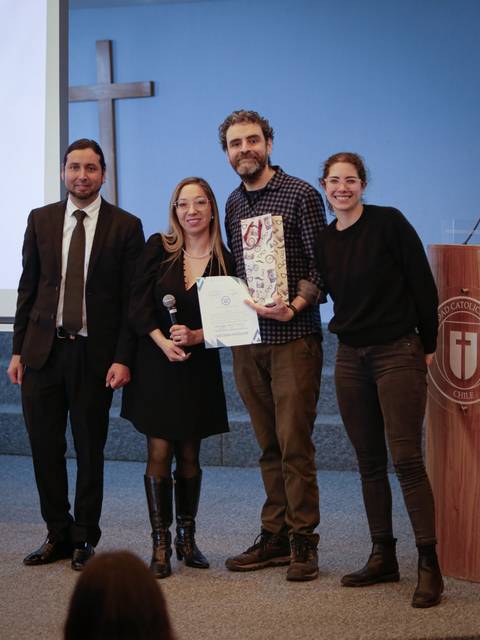 Un grupo de cuatro personas posando juntas en un evento, con un diploma en mano y un fondo institucional.