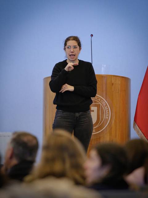 Una mujer está hablando y gesticulando frente a una audiencia en una sala.