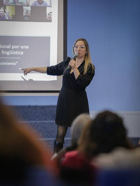 Una mujer presenta en una conferencia frente a una audiencia mientras señala hacia una pantalla con contenido digital.