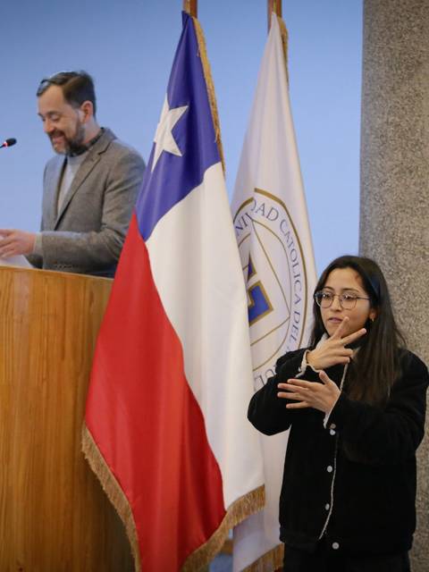 Una ceremonia con un orador y una intérprete de lenguaje de señas en un escenario con banderas.