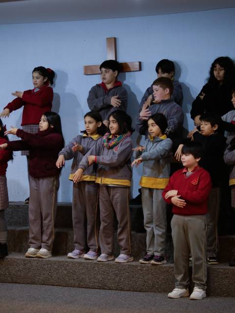 Un grupo de niños vestidos con uniformes escolares realizan una coreografía en un escenario frente a una cruz.